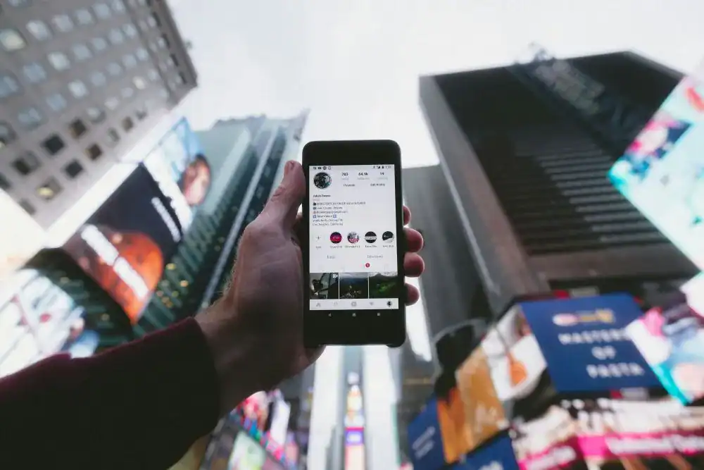 Pessoa a segurar smartphone com perfil de Instagram aberto no ecrã em ambiente urbano com arranha-céus e outdoors